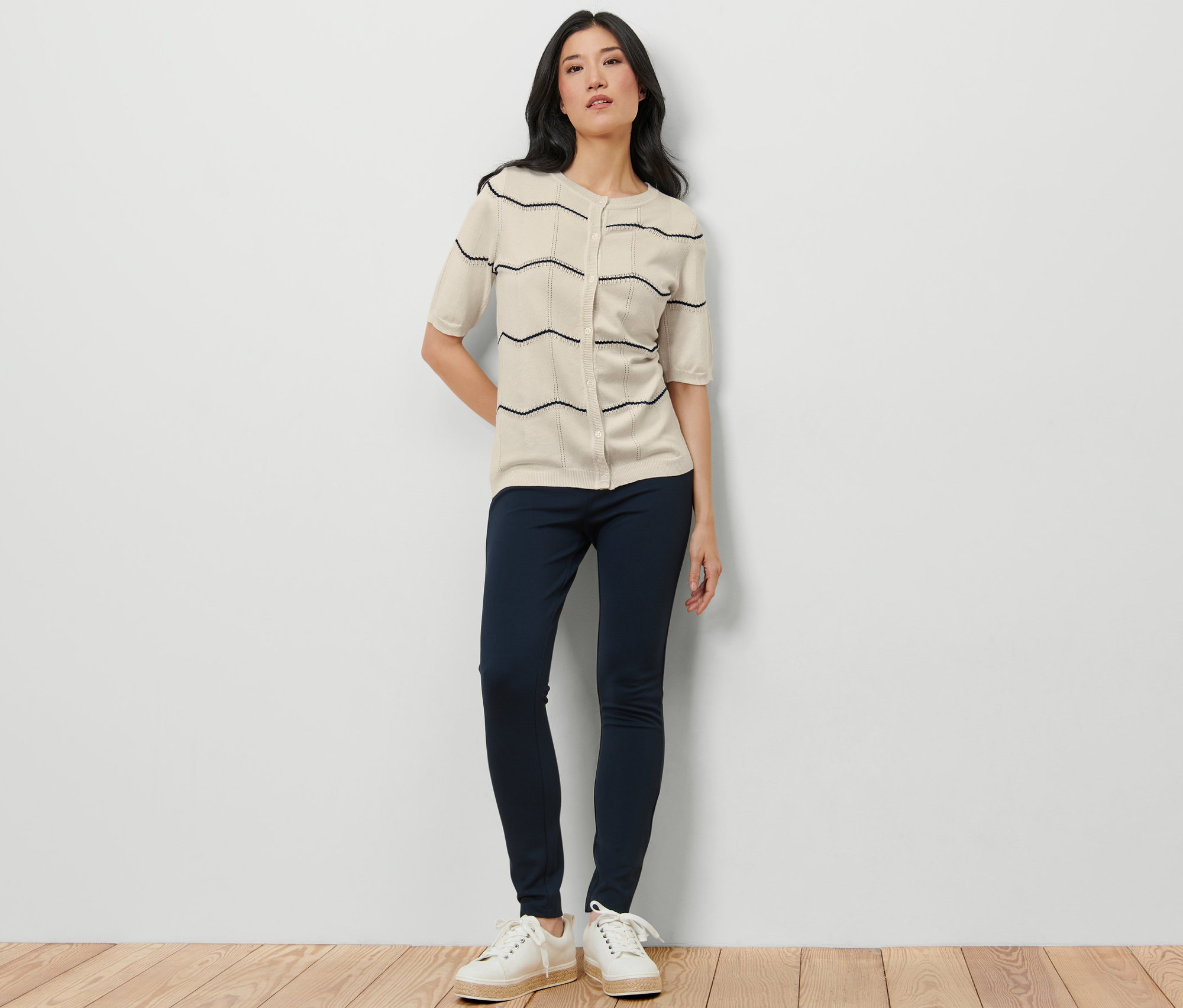 Frau steht und trägt einen Feinstrick-Cardigan mit Ajour-Muster, dunkelblaue Hosen und weiße Turnschuhe.