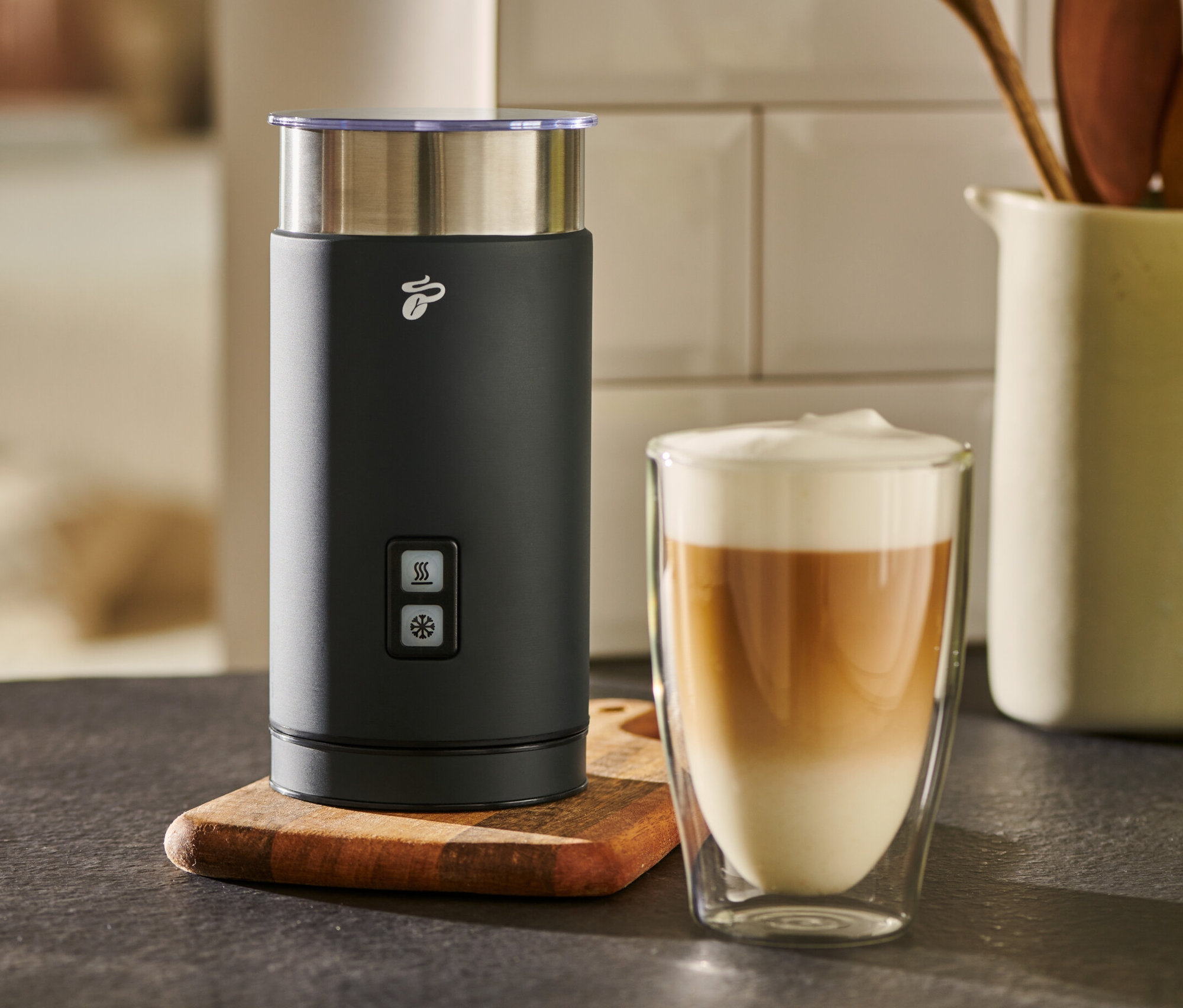 Ein schwarzer elektrischer Milchaufschäumer steht neben einem Glas Milchkaffee.