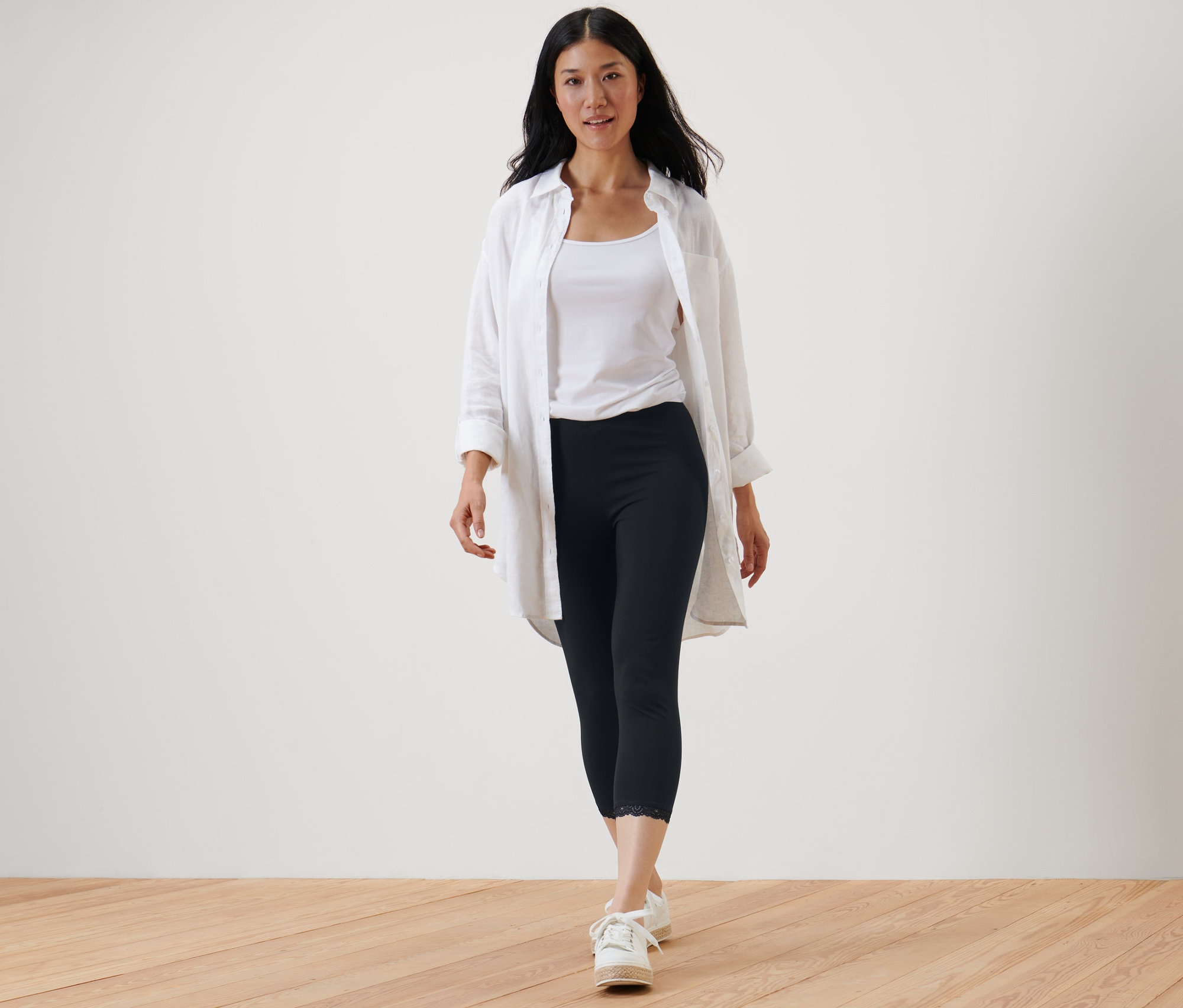 Frau geht und trägt eine schwarze 3/4-Leggings mit Spitzenborte, ein weißes Tanktop und ein weißes Hemd.