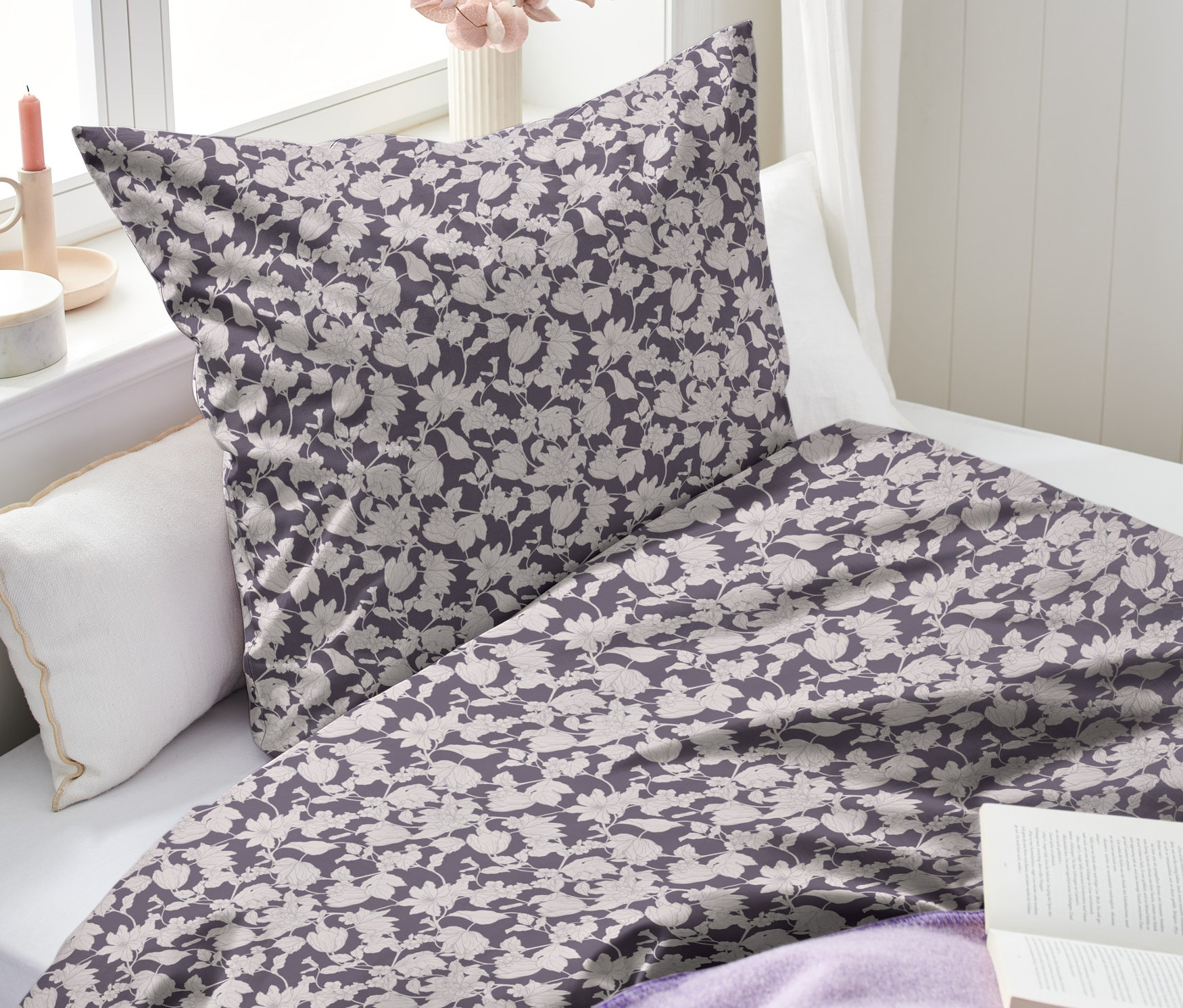 Lila Renforcé-Bettwäsche mit Blumenmuster liegt auf einem Bett. Daneben liegen ein offenes Buch und eine lila Decke.