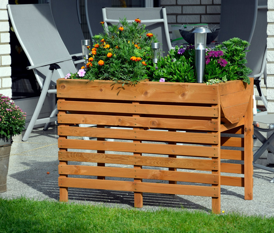Ein braunes, doppeltes XL-Hochbeet mit Pflanzen steht auf einer Terrasse neben grauen Stühlen.