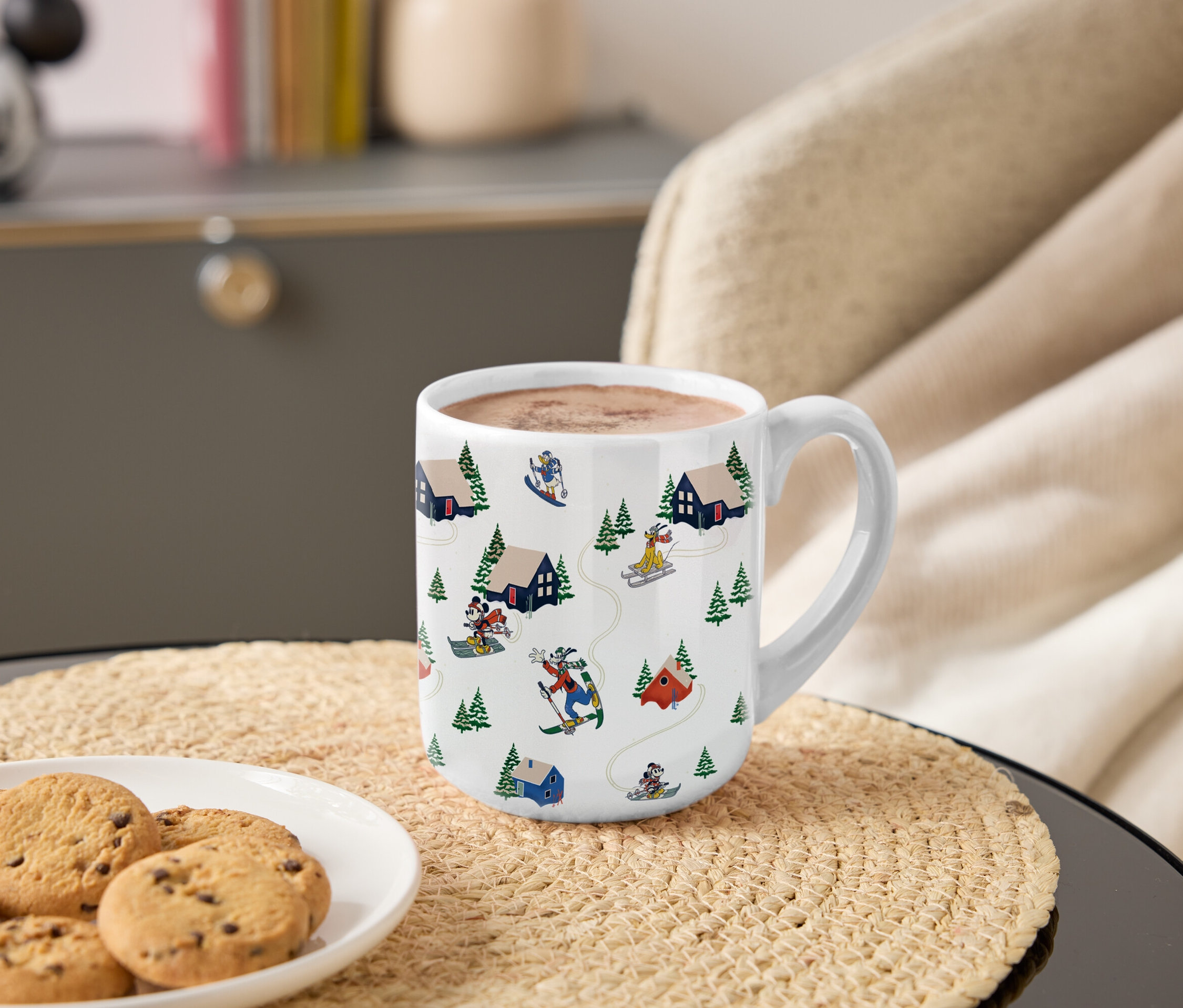 Eine Tasse mit einem Motiv von Mickey und seinen Freunden beim Skifahren in den Bergen steht auf einem Tisch neben einem Teller mit Keksen.