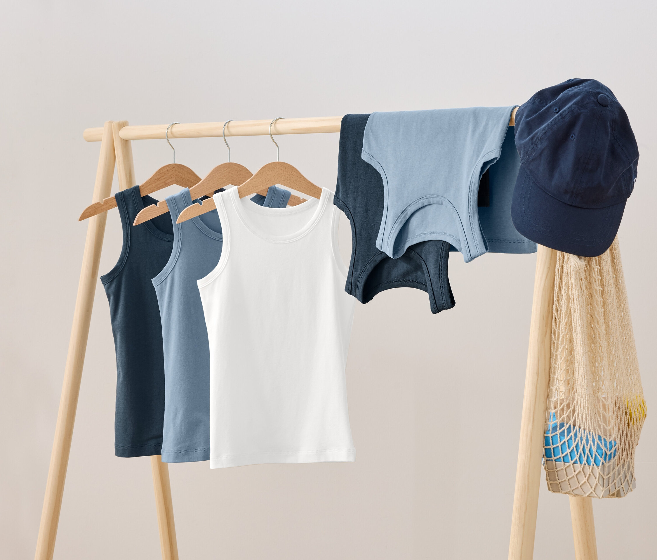 Auf einem hölzernen Kleiderständer hängen drei Tanktops auf Bügeln, daneben liegen zwei weitere Tanktops, eine blaue Schirmmütze und eine Netztasche.