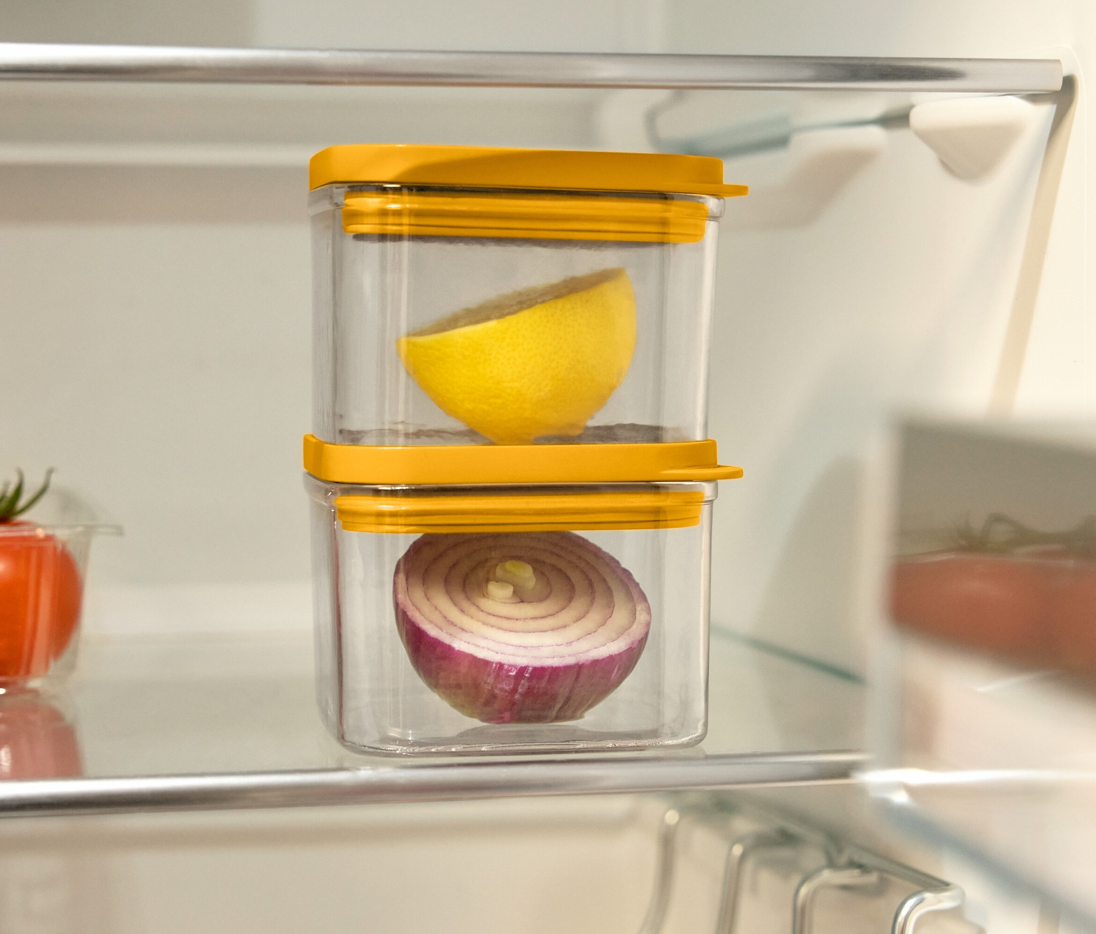Im Kühlschrank stehen zwei durchsichtige Behälter mit gelben Deckeln übereinander. Im oberen Behälter befindet sich eine halbe Zitrone und im unteren eine halbe rote Zwiebel. Links davon befindet sich ein Plastikbehälter mit Tomaten.