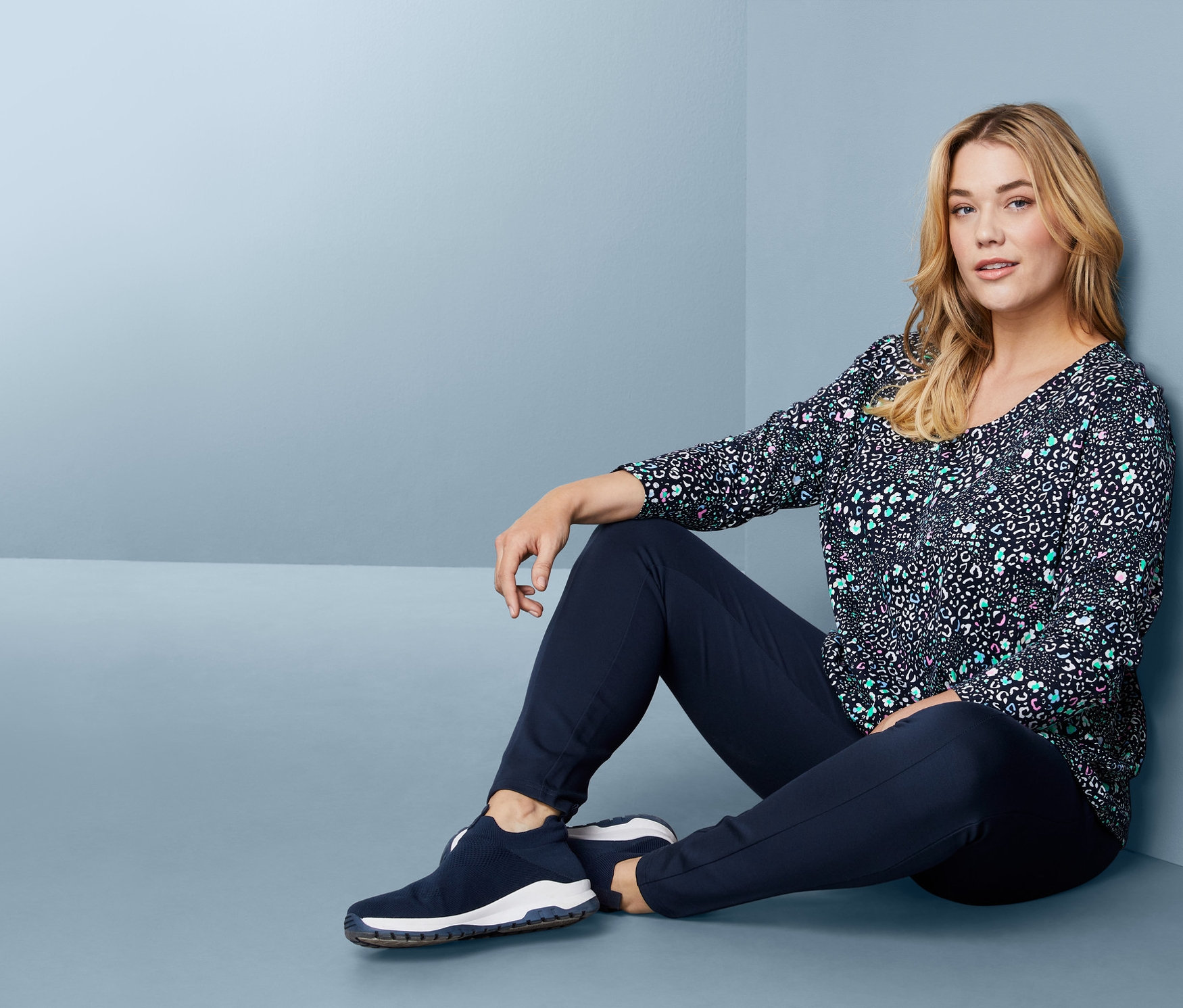 Frau sitzt auf dem Boden und trägt ein Langarmshirt, eine Bodyforming-Jerseyhose und Slip-on-Sneaker.