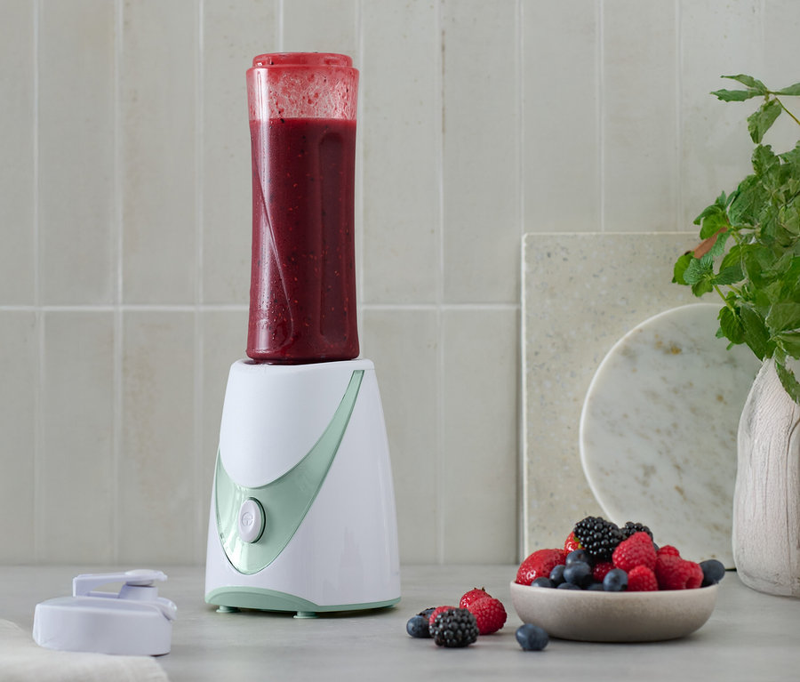 Ein Mini-Smoothie-Maker mit einem fertigen Smoothie darauf, neben einer Schale mit Beeren.