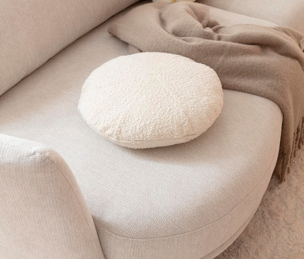 Detail eines beigen Sofas mit einem runden, weißen Kissen, einem gelb gemusterten Kissen und einer beigen Decke.