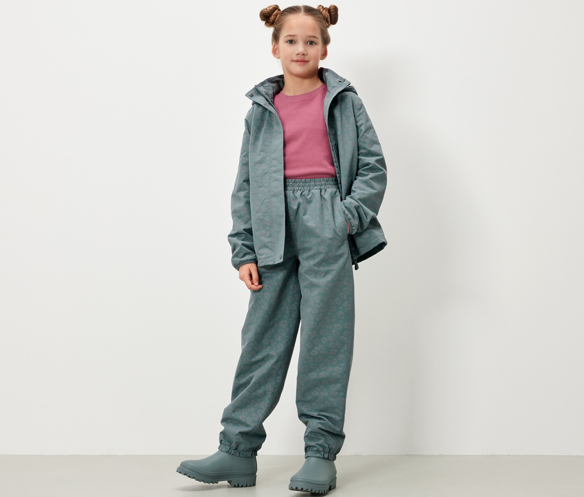 Mädchen posiert in Kinder-Regenhose mit reflektierenden Elementen, Jacke und Gummistiefeln in Graugrün.