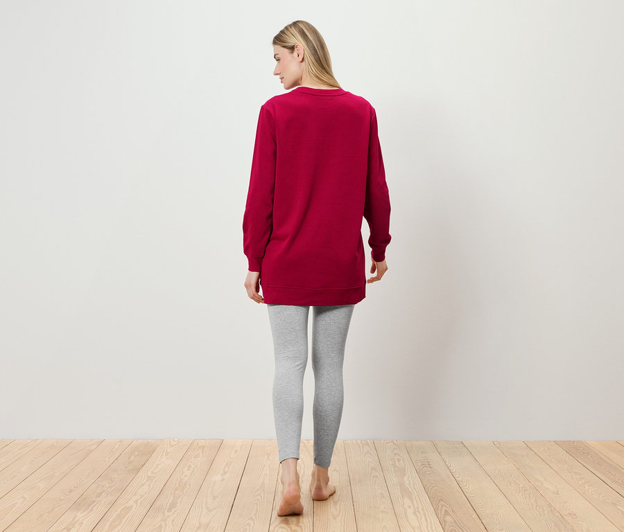 Eine Frau steht mit dem Rücken zur Kamera und trägt eine weinrote Tunika und graue Leggings.