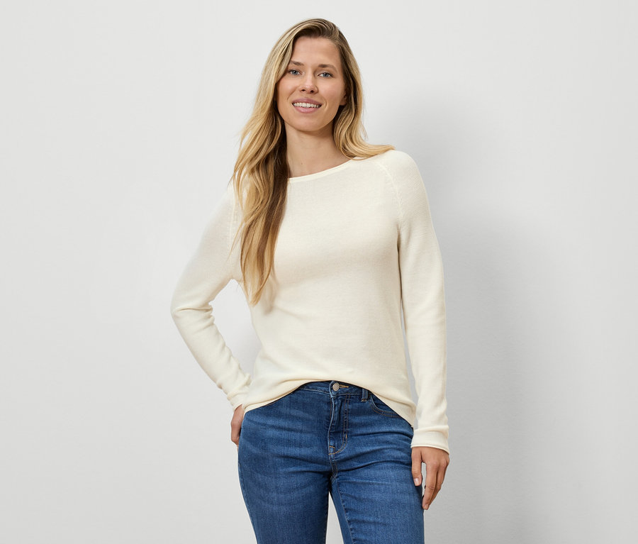 Frau mit blonden Haaren posiert in einem cremefarbenen Pullover und blauen Jeans.