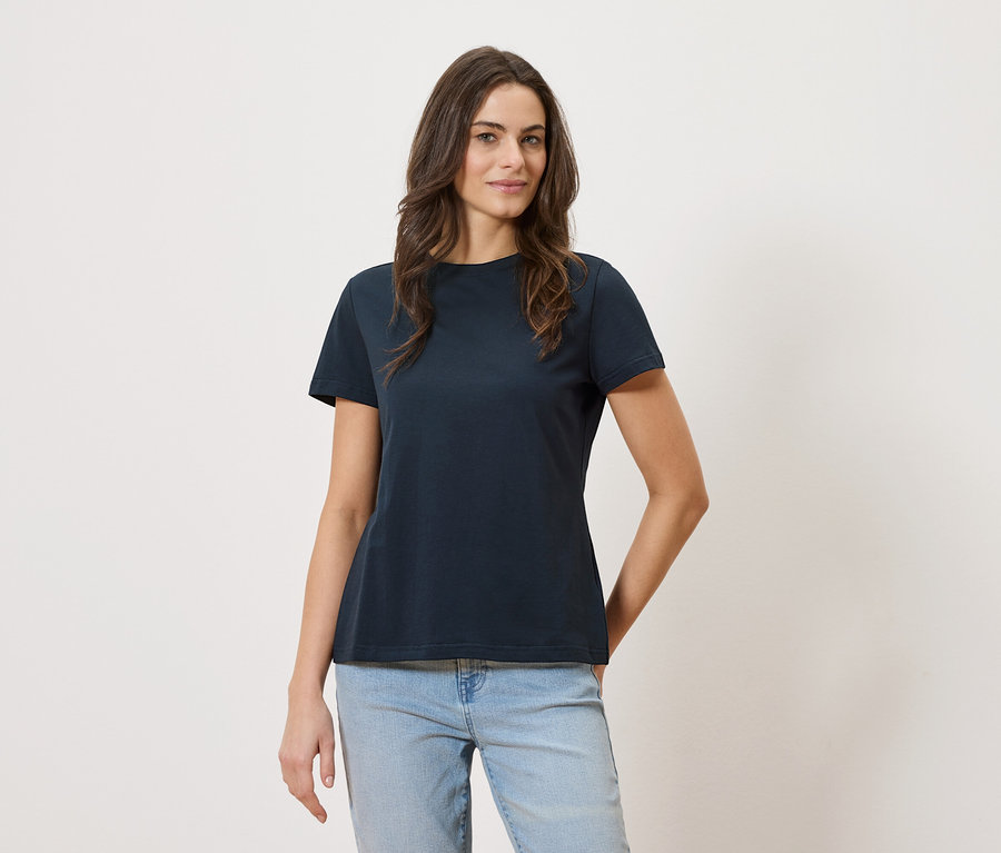 Frau trägt ein marineblaues T-Shirt und Jeans vor einem weißen Hintergrund.