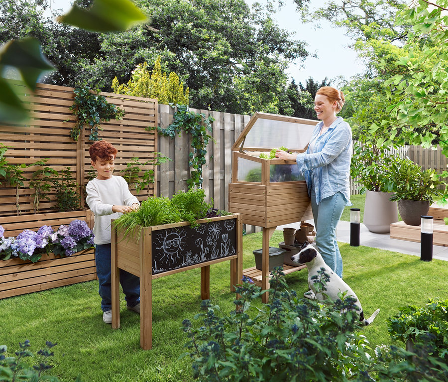 Eine Frau öffnet ein Frühbeet, während ein Kind neben einem Hochbeet steht. Ein Hund sitzt im Gras.
