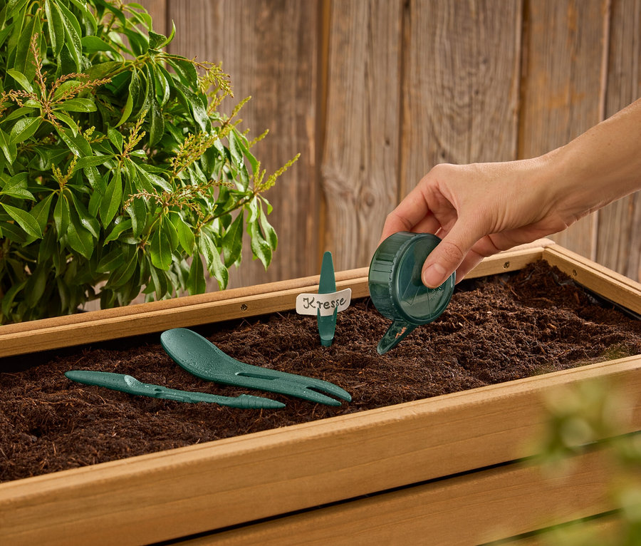 Eine Hand gießt Wasser aus einer kleinen grünen Gießkanne in einen Holzkasten mit Erde, in dem grüne Gartengeräte und ein Kresseschild stecken.