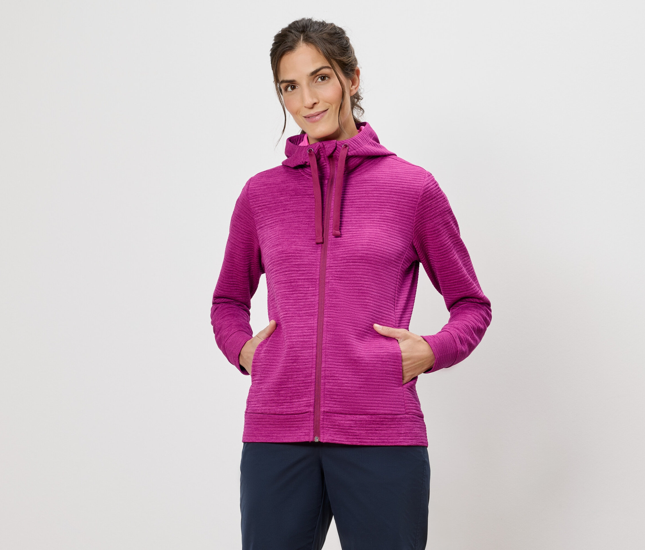 Frau mit Kapuzenpullover in Pink mit Händen in den Taschen vor einem weißen Hintergrund.