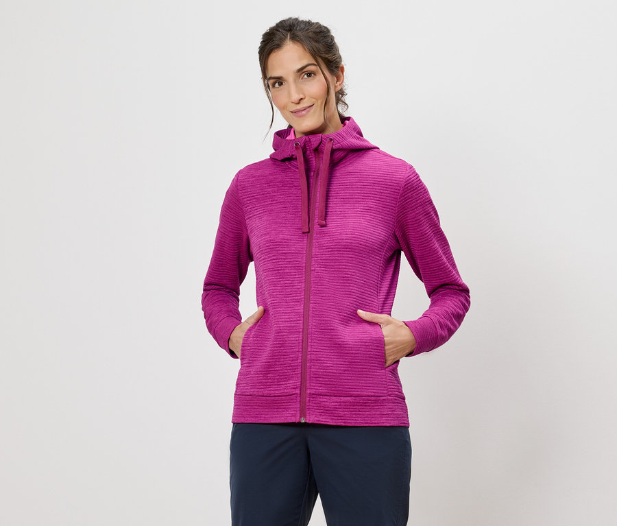 Frau mit Kapuzenpullover in Pink mit Händen in den Taschen vor einem weißen Hintergrund.