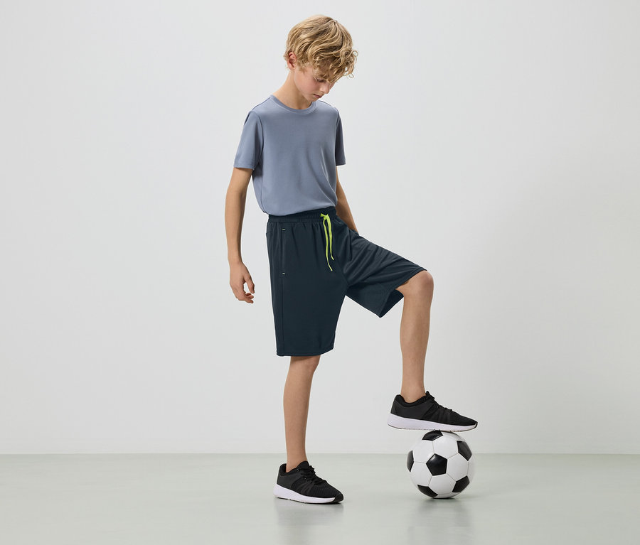 Ein blonder Junge in einem grauen T-Shirt und dunklen Shorts mit einem Fuß auf einem Fußball.