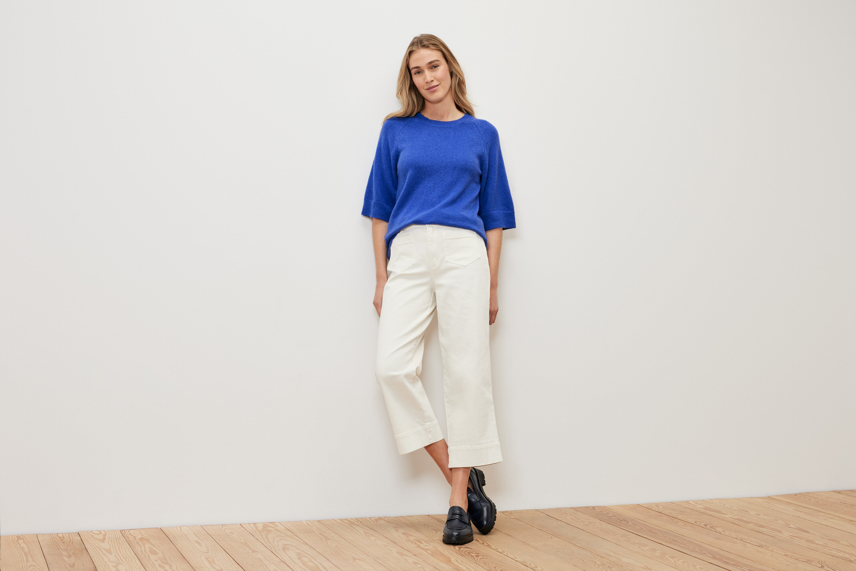 Eine Frau steht vor einer weißen Wand und trägt einen kobaltblauen Strickpullover, weiße Hosen und schwarze Schuhe.