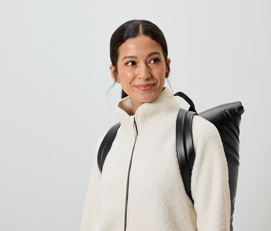 Lächelnde Frau mit braunen Haaren und braunen Augen trägt eine cremefarbene Jacke und einen schwarzen Rucksack.