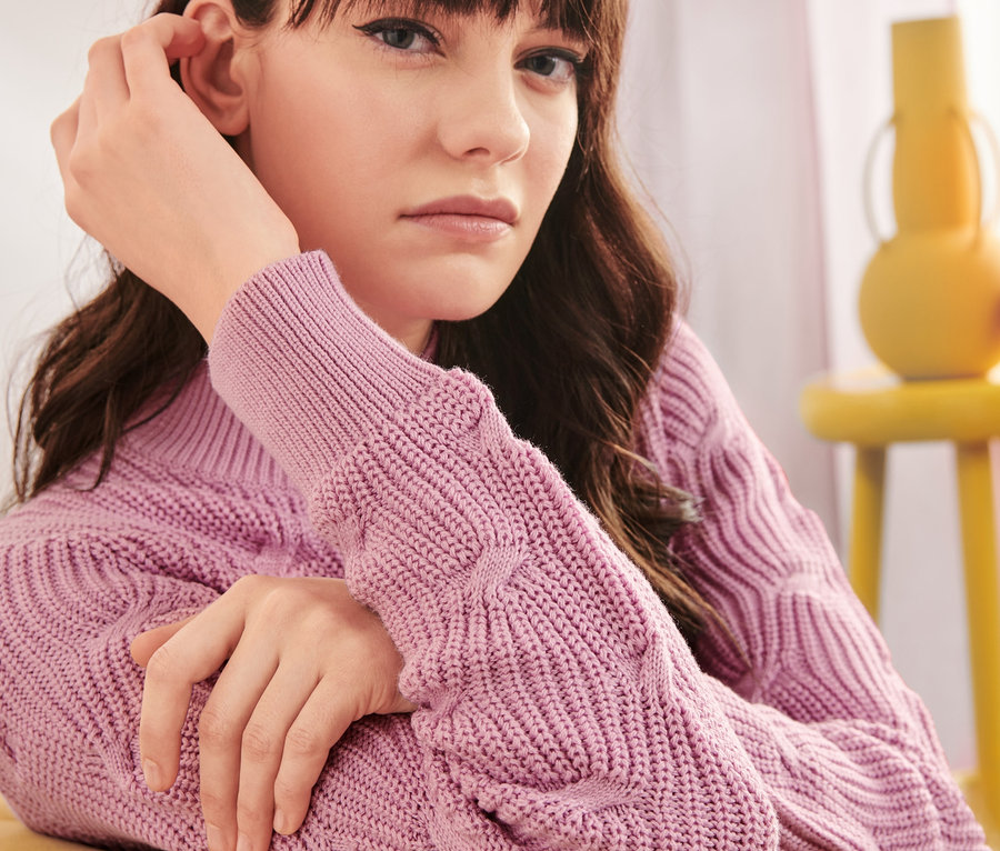 Frau sitzt und trägt einen Pullover mit Zopfmuster.