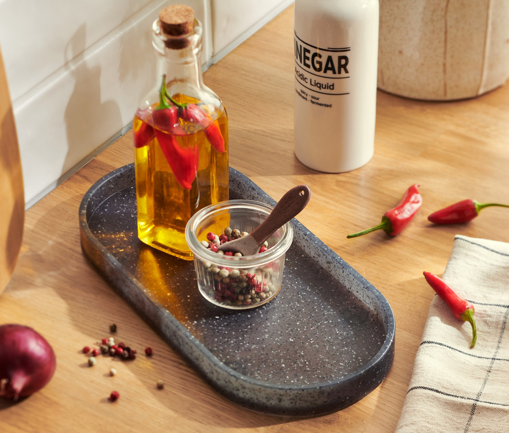 Auf einer Küchentheke steht eine Küchenablage in Granitoptik mit einer Ölflasche, Chilischoten und einem Gewürzglas.
