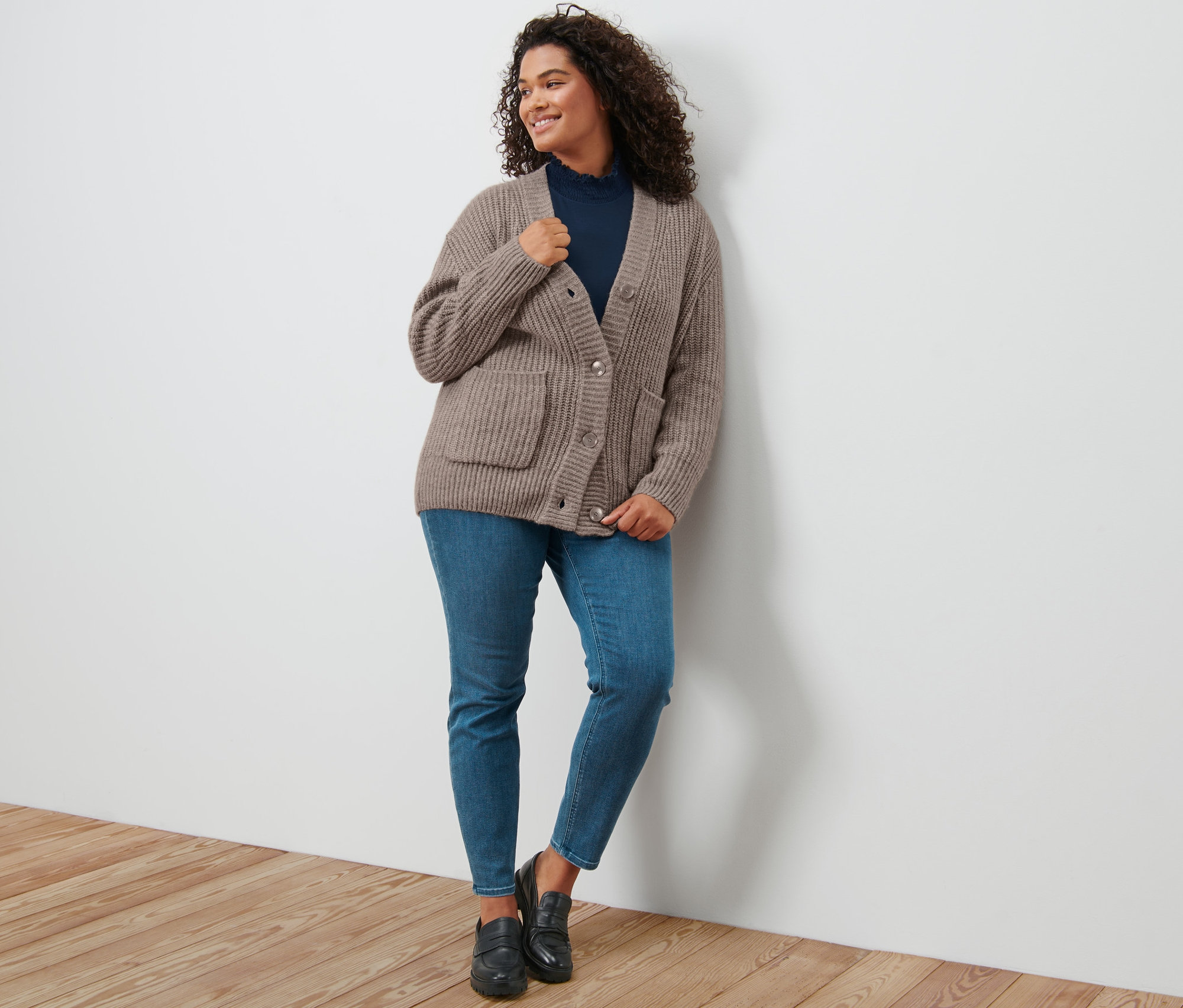 Eine Frau lehnt an einer Wand und trägt einen taupefarbenen, gerippten Grobstrick-Cardigan, ein dunkelblaues Blusenshirt mit Stehkragen, Treggings »Fit Nelly« und Leder-Loafer.