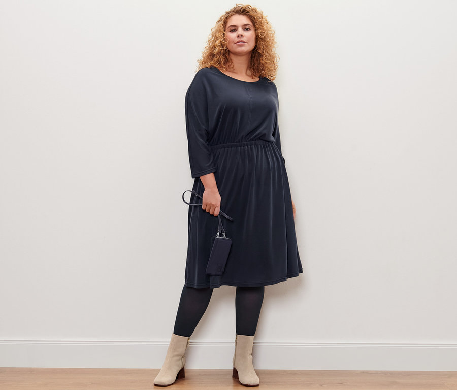 Frau posiert in einem Piqué-Jerseykleid und hält eine Mini-Tasche. Sie trägt 2 Paar Feinstrumpfhosen.