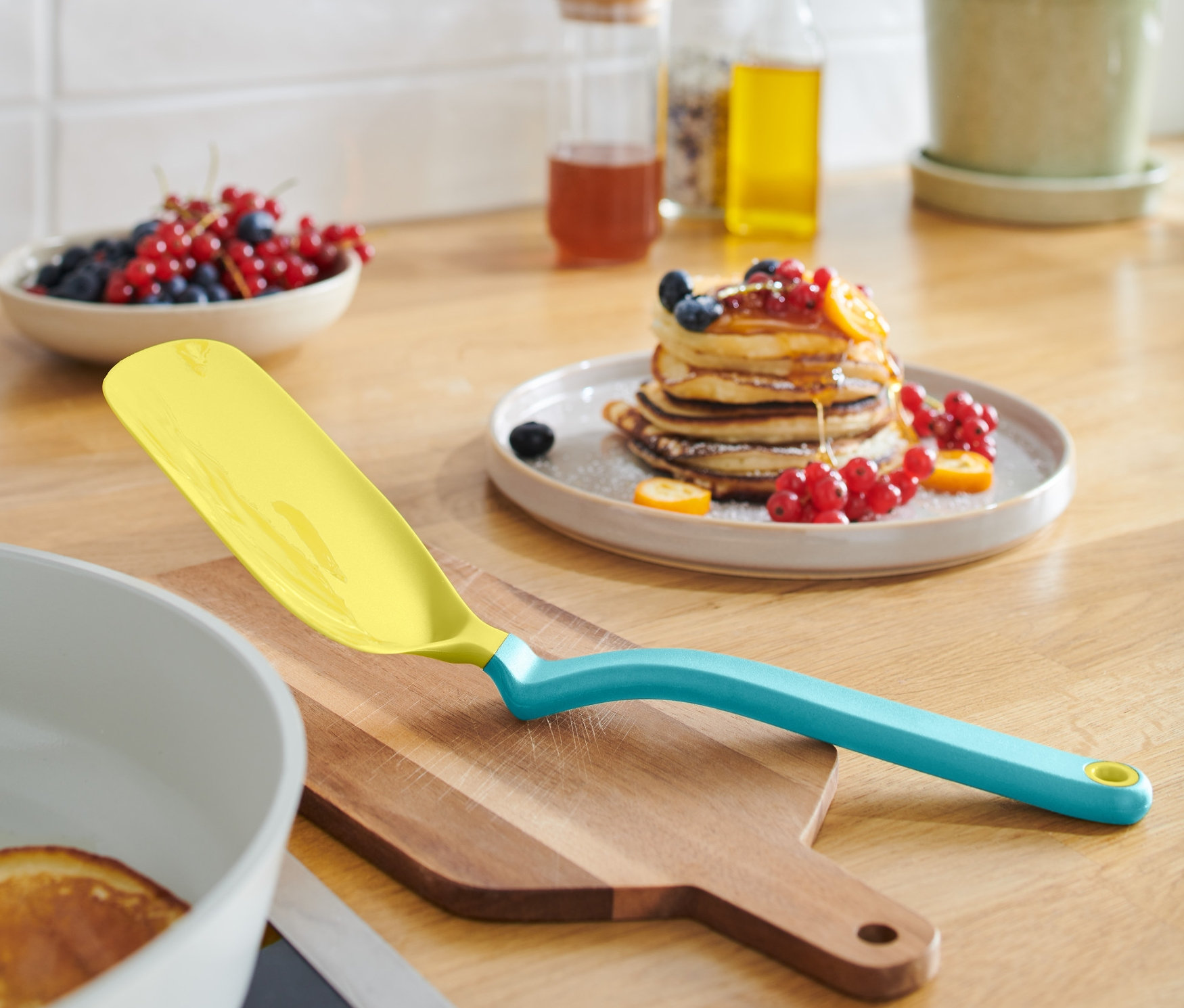 Gelber Eier-und-Pfannkuchenwender liegt auf einem Holzbrett neben einer Pfanne mit Pfannkuchen.