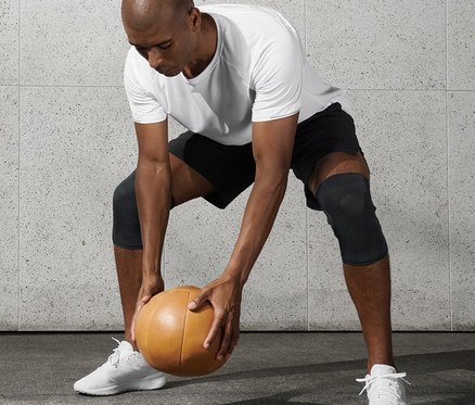 Mann in weißem T-Shirt und schwarzen Shorts hält Medizinball und trägt 2 Fitness-Knie-Manschetten.