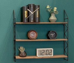 Zwei runde Wanduhren und ein Regal mit drei Ablagen an einer grünen Wand. Auf den Regalen stehen Bücher, eine Vase und verschiedene Uhren. Darunter eine Tischuhr »Buch«.