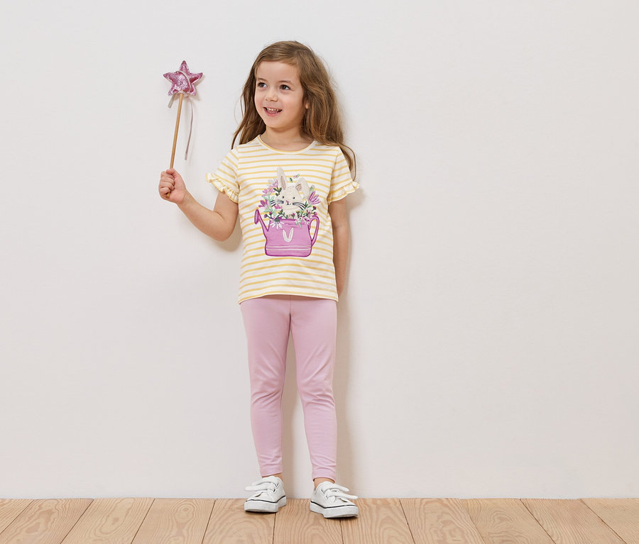 Mädchen hält einen Sternenstab und trägt ein gestreiftes T-Shirt mit einer Gießkanne und Blumen darauf, sowie eine rosafarbene Leggings.