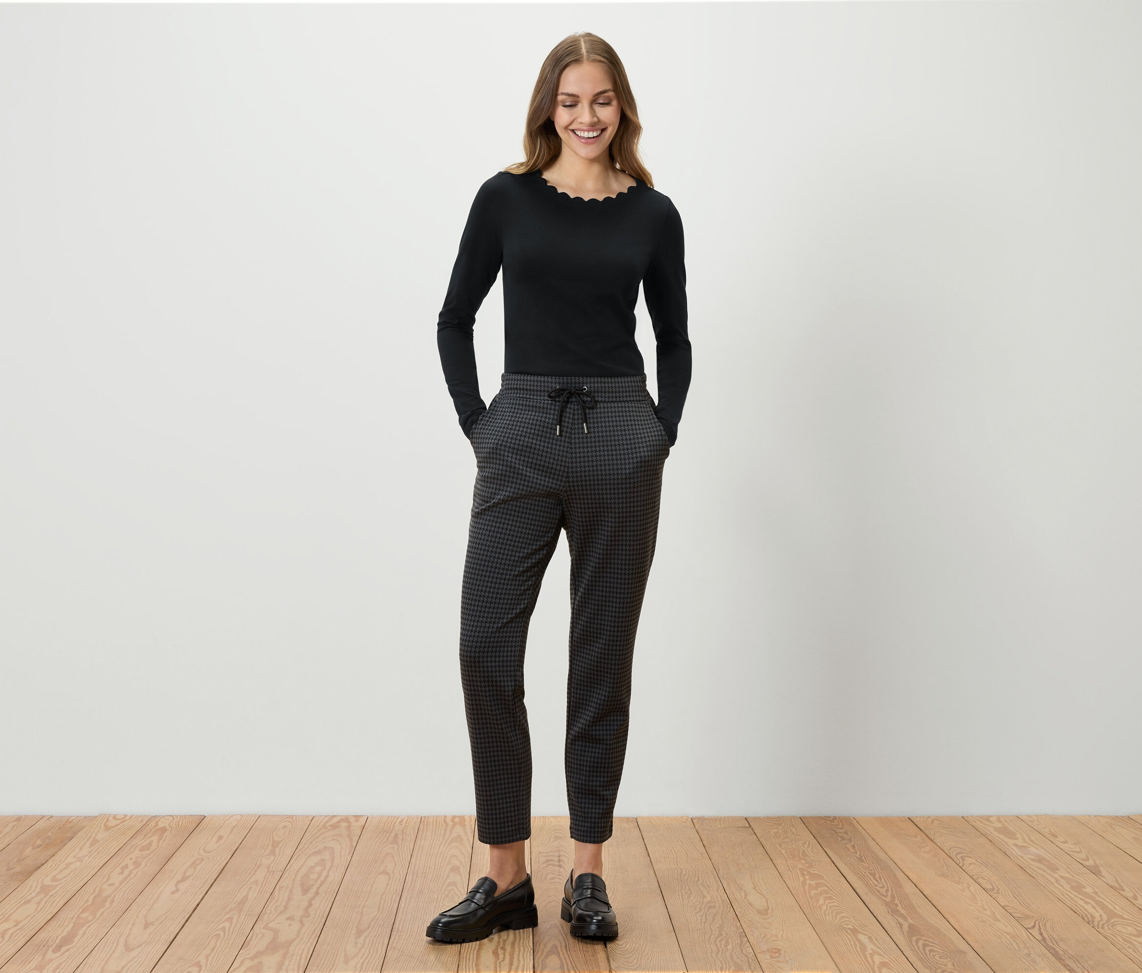 Eine Frau steht und lächelt, sie trägt ein schwarzes Langarmshirt, eine graue gemusterte Hose und schwarze Slipper.