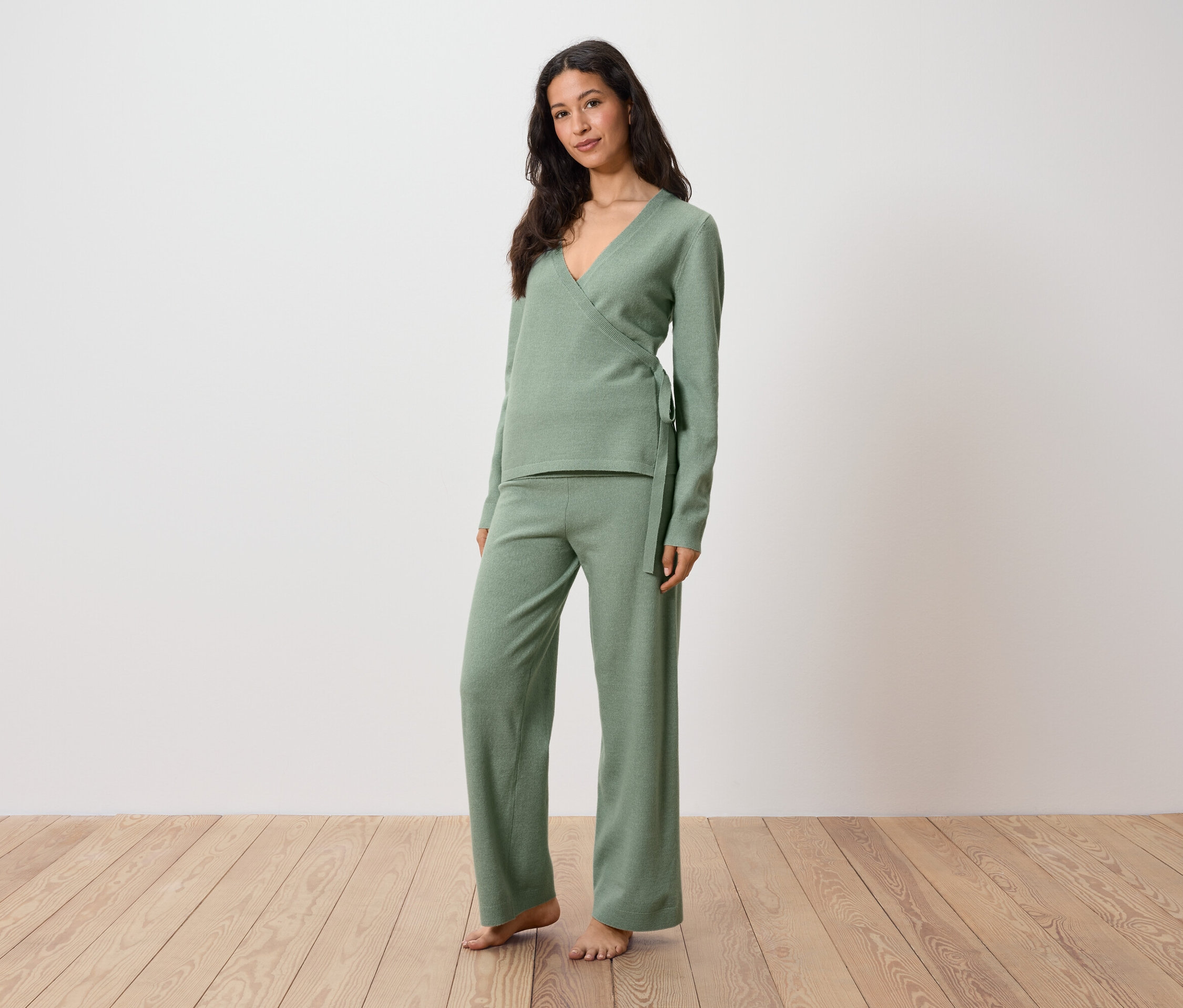 Frau in grünem Wickeloberteil und Hose steht auf Holzboden.