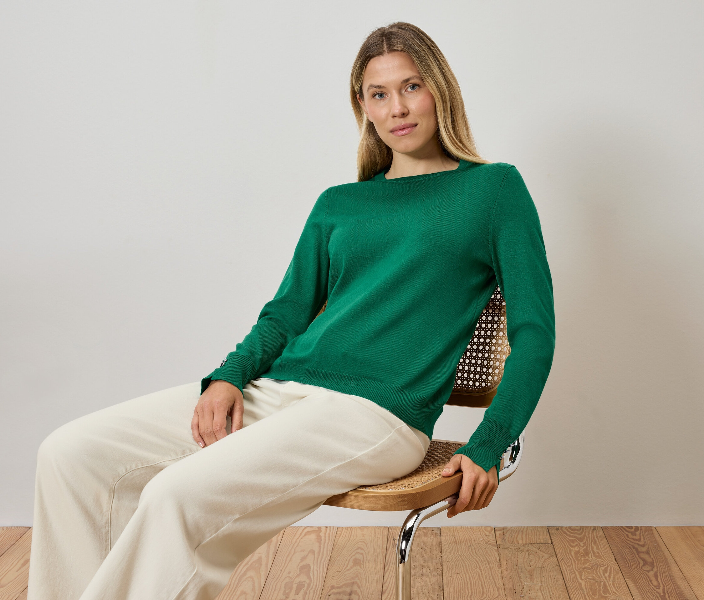 Blonde Frau sitzt auf einem Stuhl, trägt einen grünen Pullover und cremefarbene Hosen.