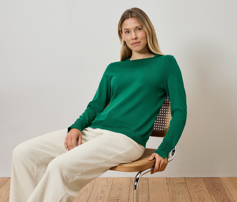 Blonde Frau sitzt auf einem Stuhl, trägt einen grünen Pullover und cremefarbene Hosen.