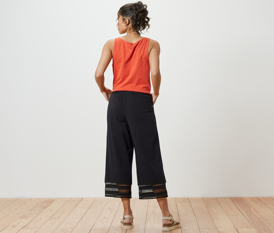 Eine Frau steht mit dem Rücken zum Betrachter und trägt ein orangefarbenes Top und eine schwarze Culotte mit Stickerei.