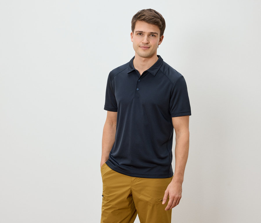 Mann mit marineblauem Poloshirt und goldbraunen Hosen, eine Hand in der Hosentasche.