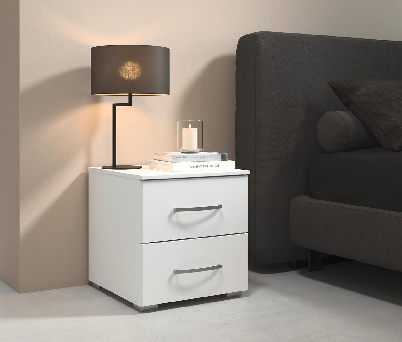 Weißer rauch Nachttisch »Aditio« mit 2 Schubladen neben einem grauen Bett mit Kissen. Auf dem Nachttisch stehen eine Lampe, Bücher und eine Kerze.