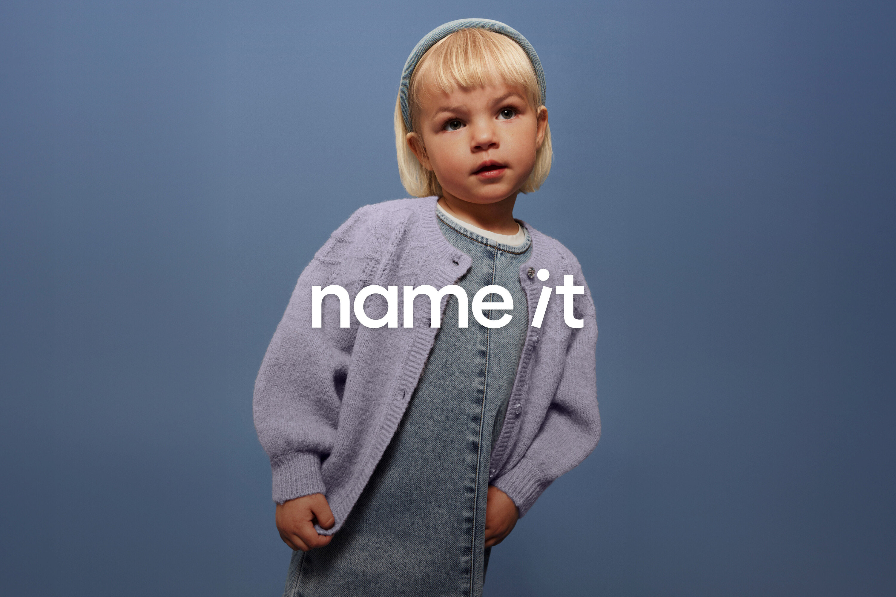 Kleines Kind mit blonden Haaren und blauem Haarband trägt eine Jeanslatzhose und eine lila Strickjacke vor einem blauen Hintergrund.