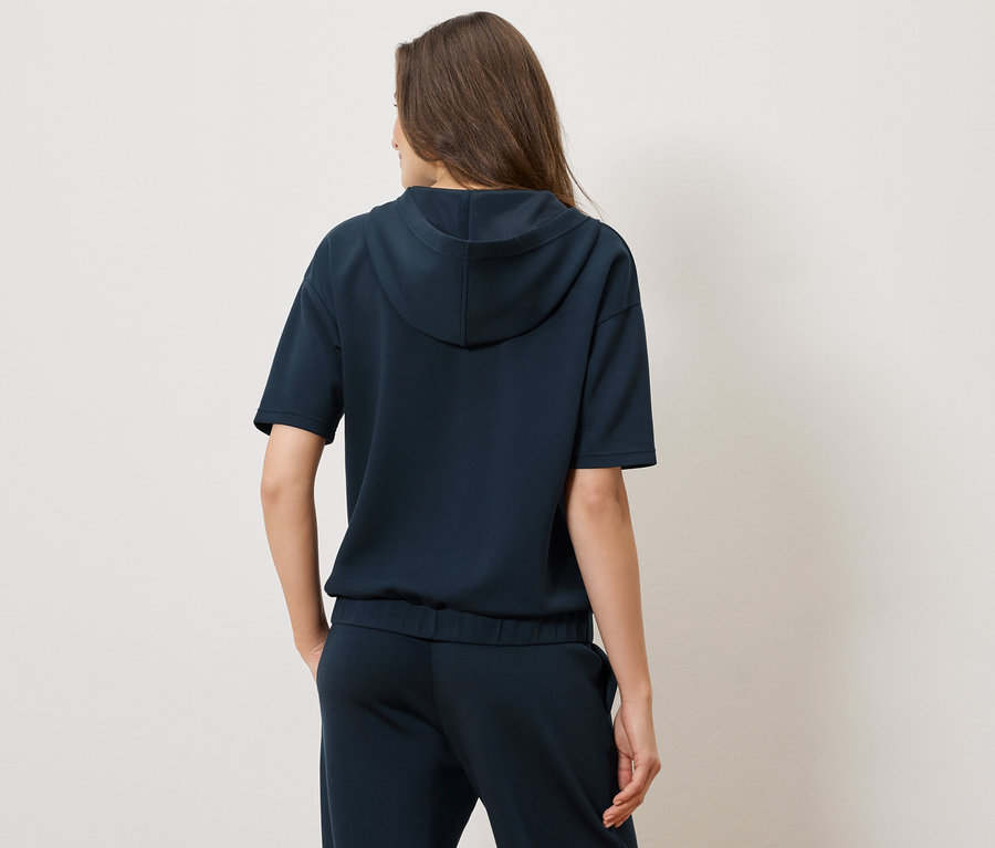 Rückansicht einer Frau mit dunkelblauem Kapuzenpulli mit kurzen Ärmeln und passender Hose.