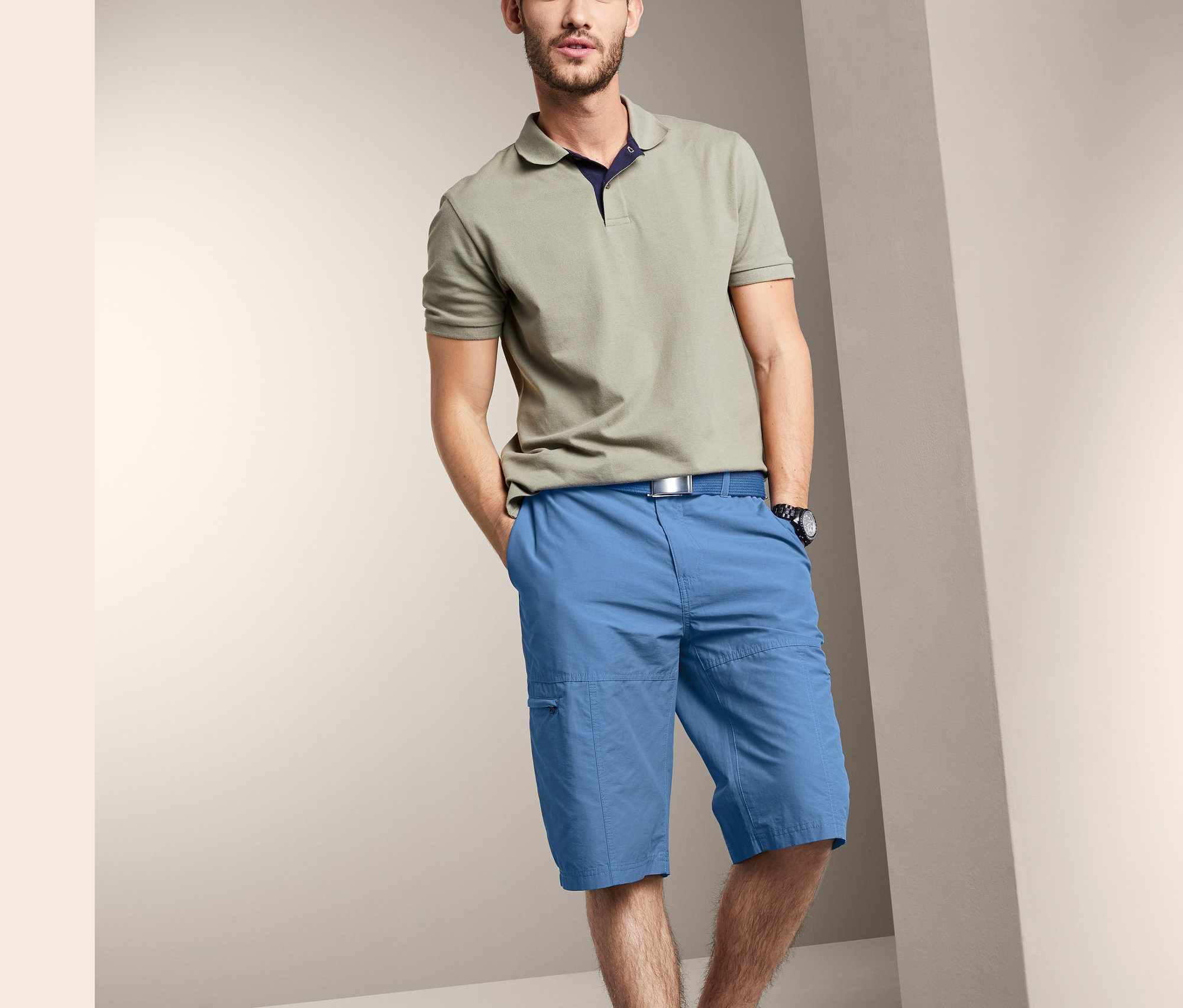 Mann posiert in einem beigen Poloshirt und blauen Bermudas.