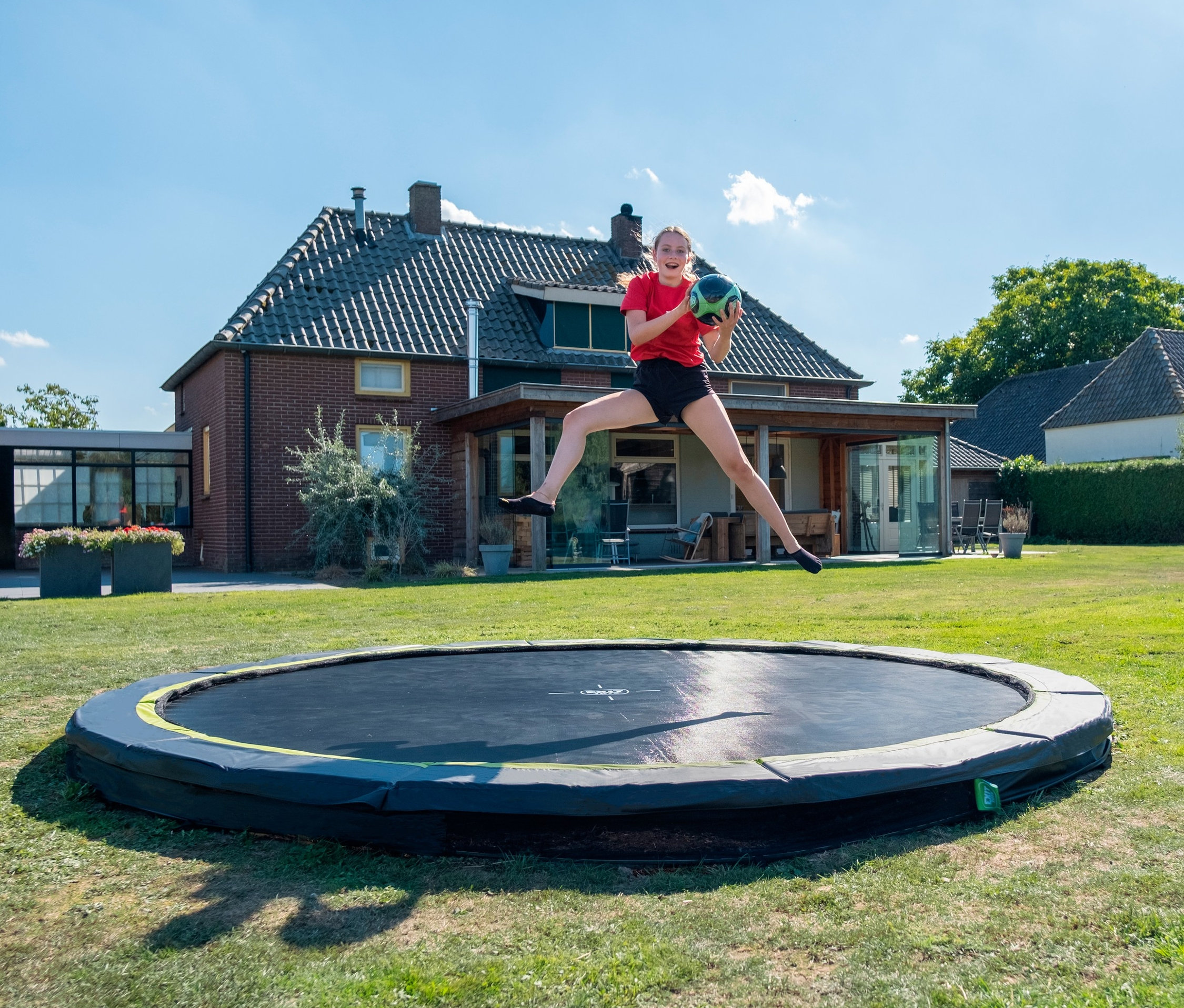 Ein junges Mädchen springt auf einem EXIT Bodentrampolin »Silhouette sports«, Ø ca. 305 cm und hält einen grünen Ball vor einem Haus.