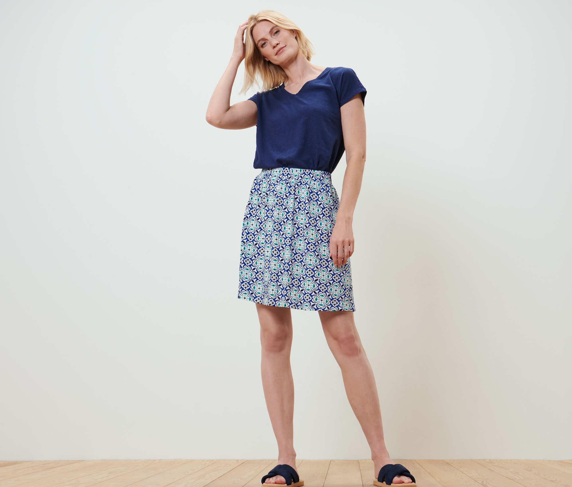 Blonde Frau steht mit der Hand im Haar. Sie trägt ein marineblaues T-Shirt und einen Jerseyrock mit Alloverprint.