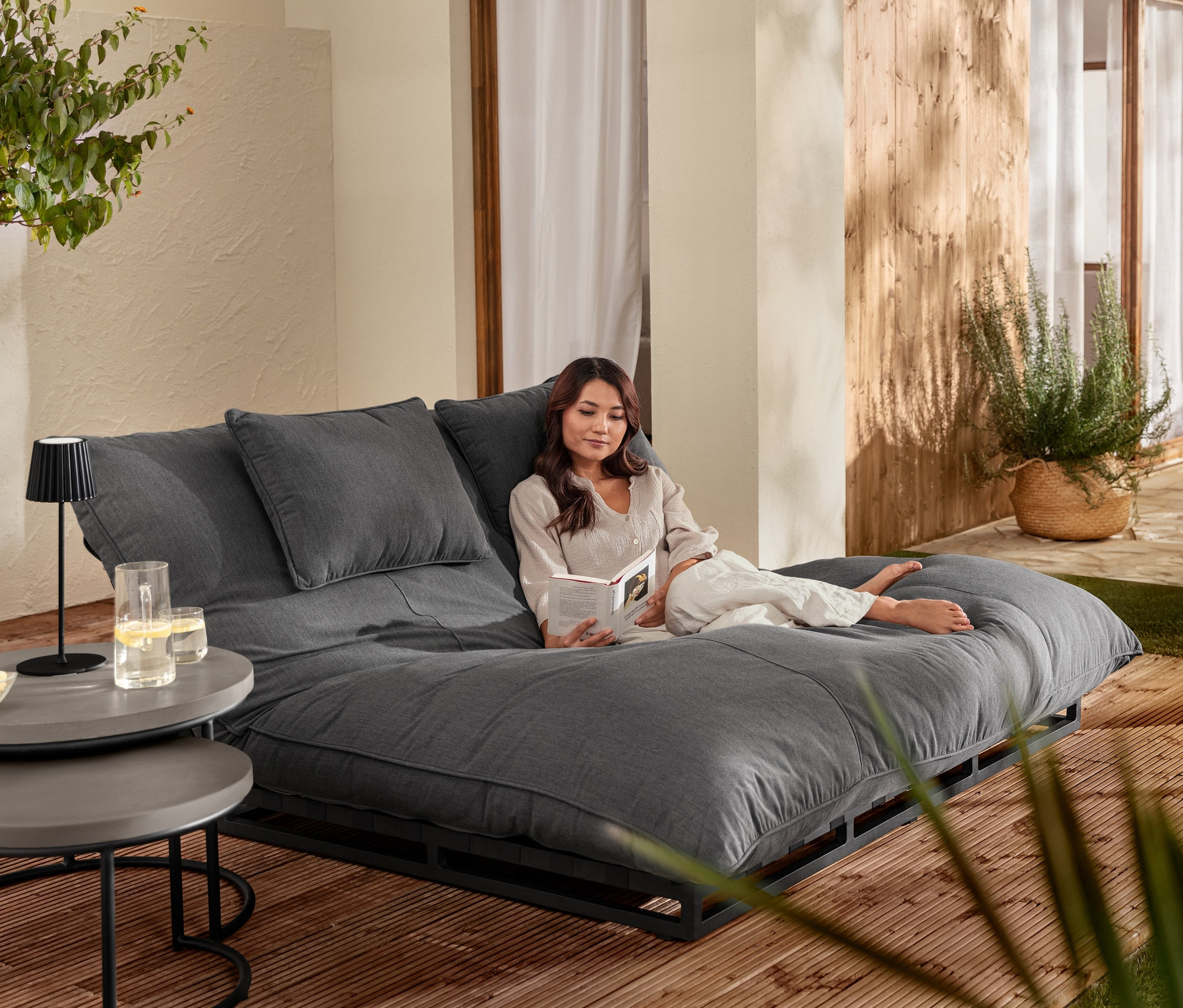 Eine Frau liest auf einer Premium-Doppelliege mit verstellbarem Kopfteil. Im Vordergrund stehen 2 Couchtische mit Beton-Tischplatte, Solar-Kugelleuchten »Granit« und eine schwarze LED-Akkutischleuchte.