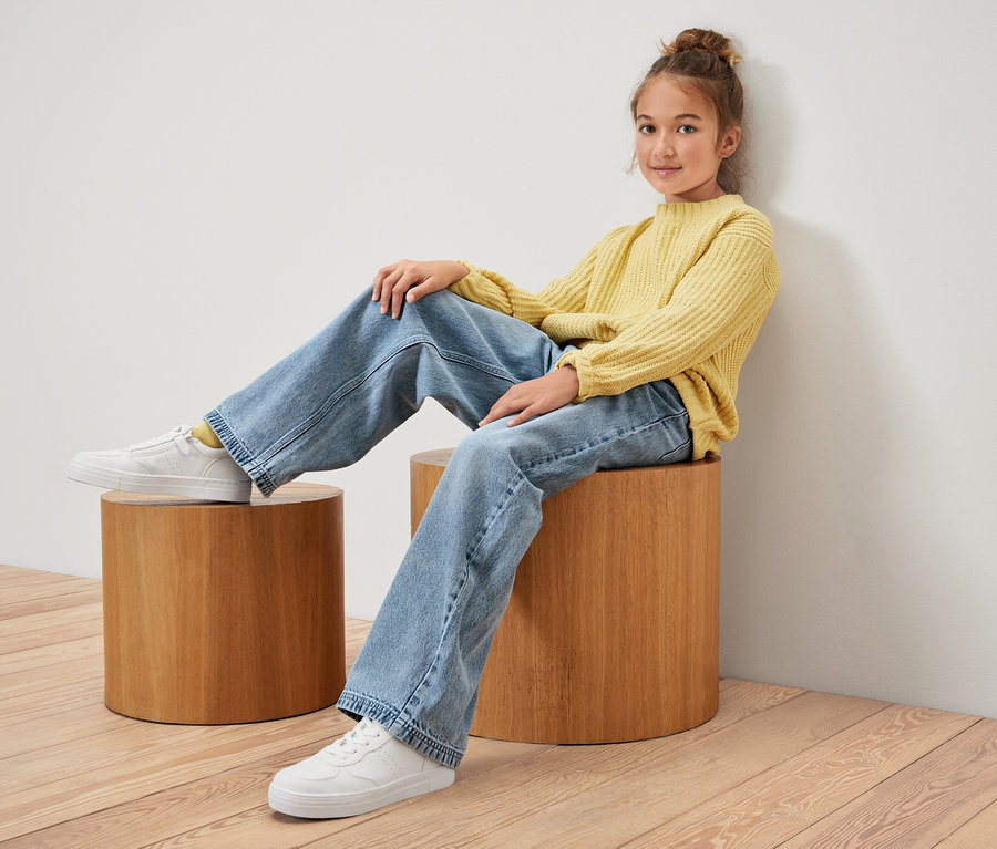 Mädchen sitzt auf einem Holzhocker und trägt einen gelben Pullover und Kinder-Jeans ‒ Fit »Lena«.