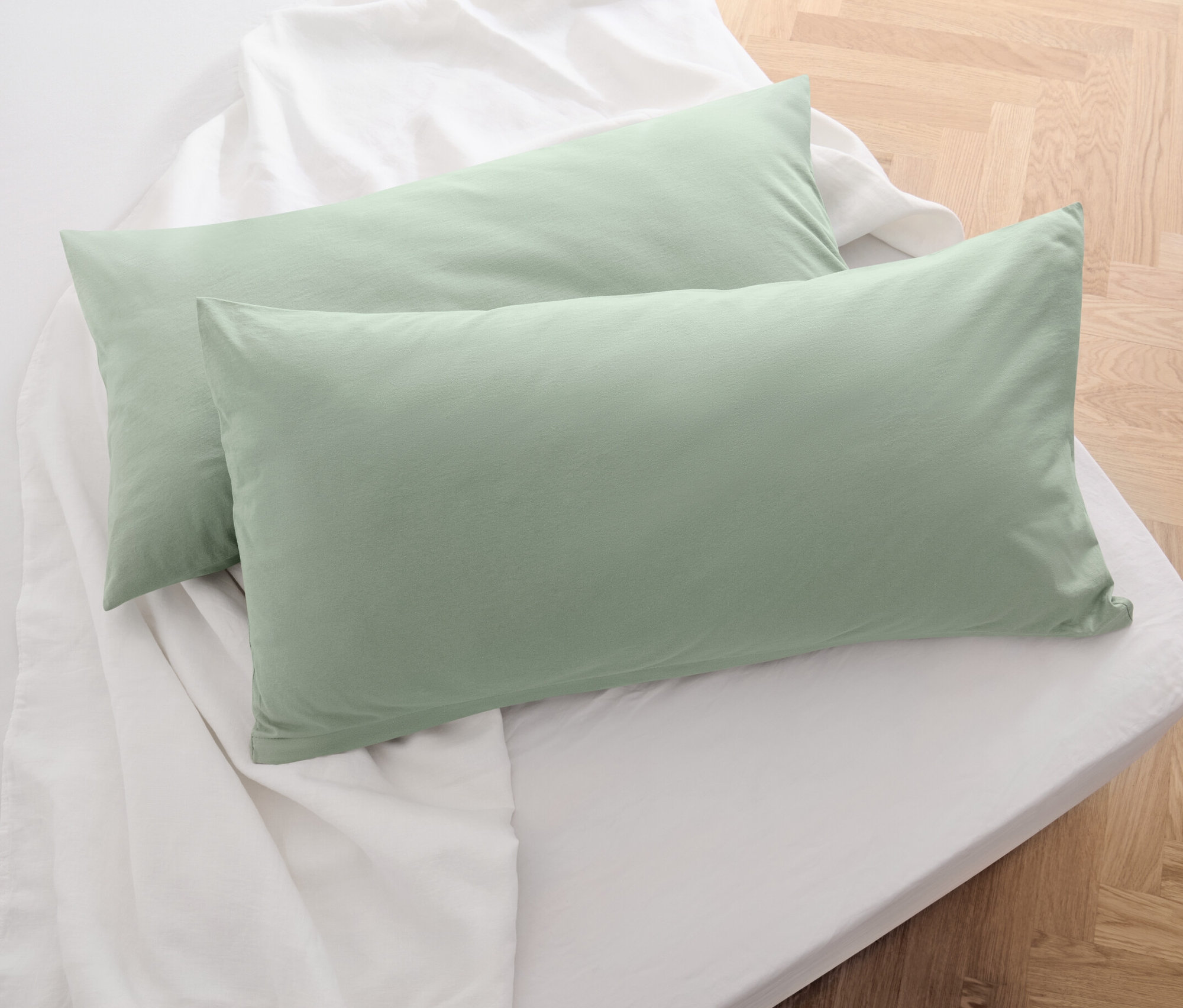 Zwei salbeigrüne Jersey-Kopfkissenbezüge liegen auf einem weißen Bettlaken.