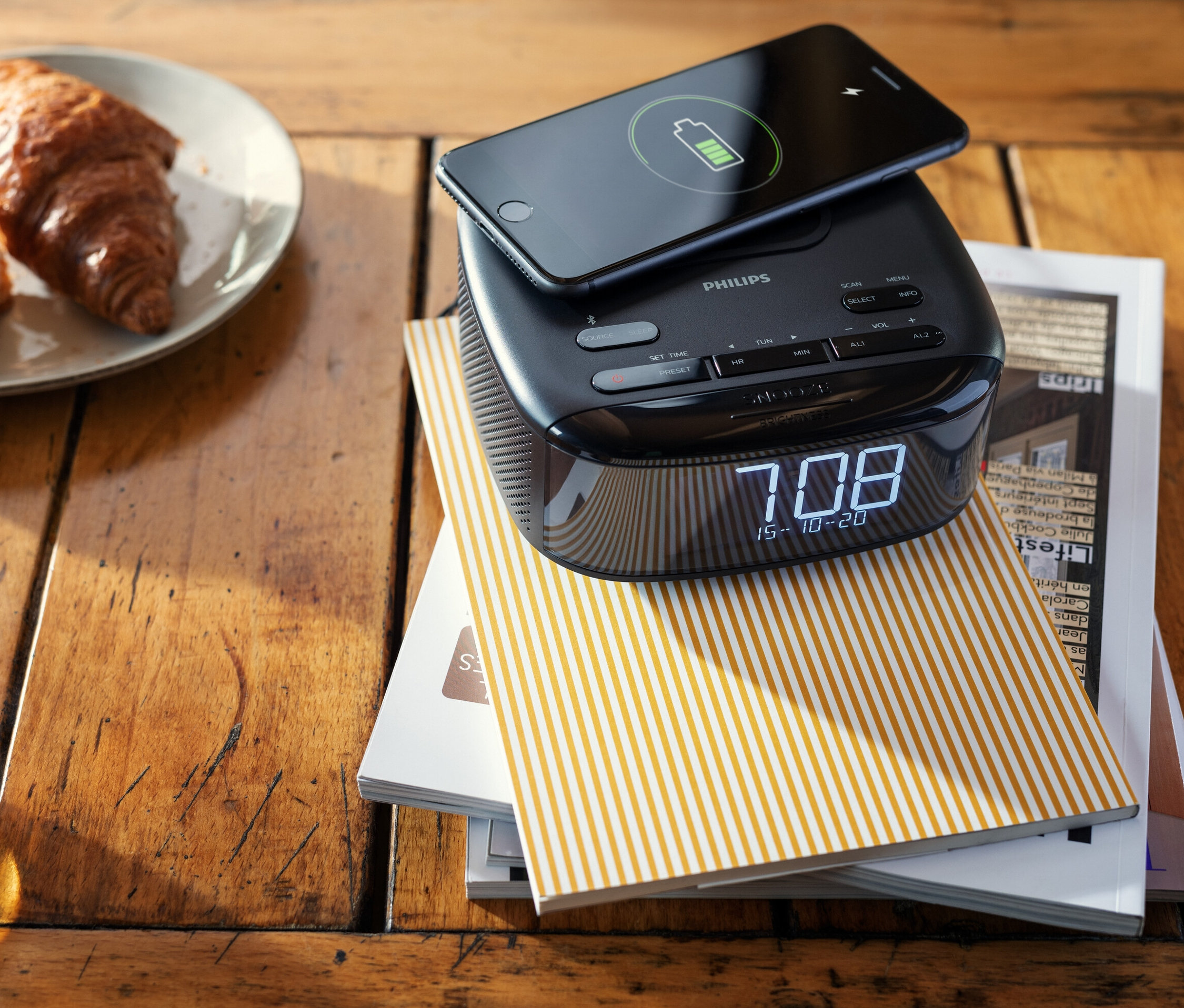 Auf einem Holztisch liegen ein Philips DAB-Radiowecker »TAR7705« mit einem aufladenden Telefon darauf, neben einem Teller mit Croissants und einer Tasse Tee.