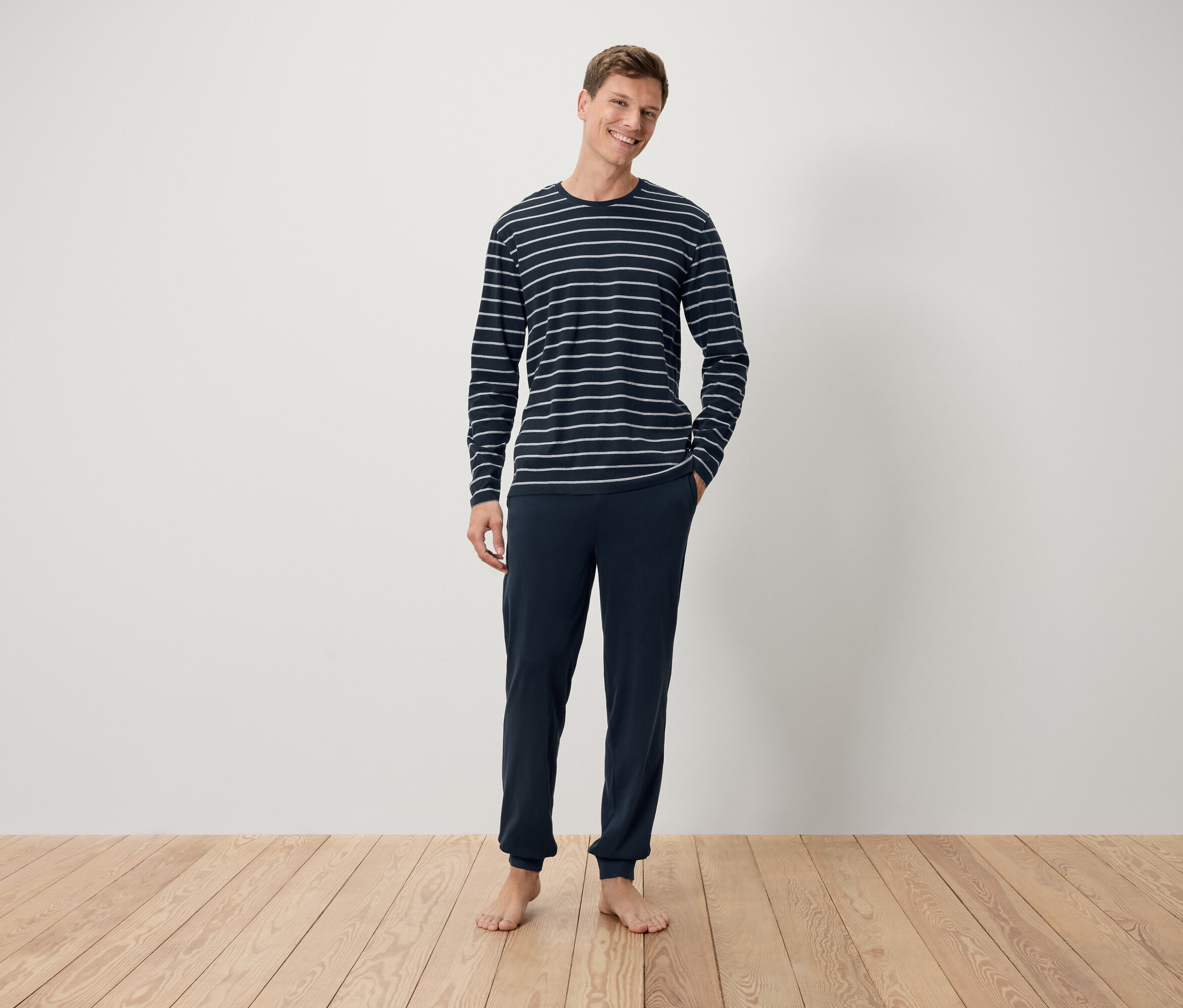 Mann steht barfuß auf einem Holzboden und trägt ein dunkelblaues, gestreiftes Langarmshirt und eine dunkelblaue Hose.