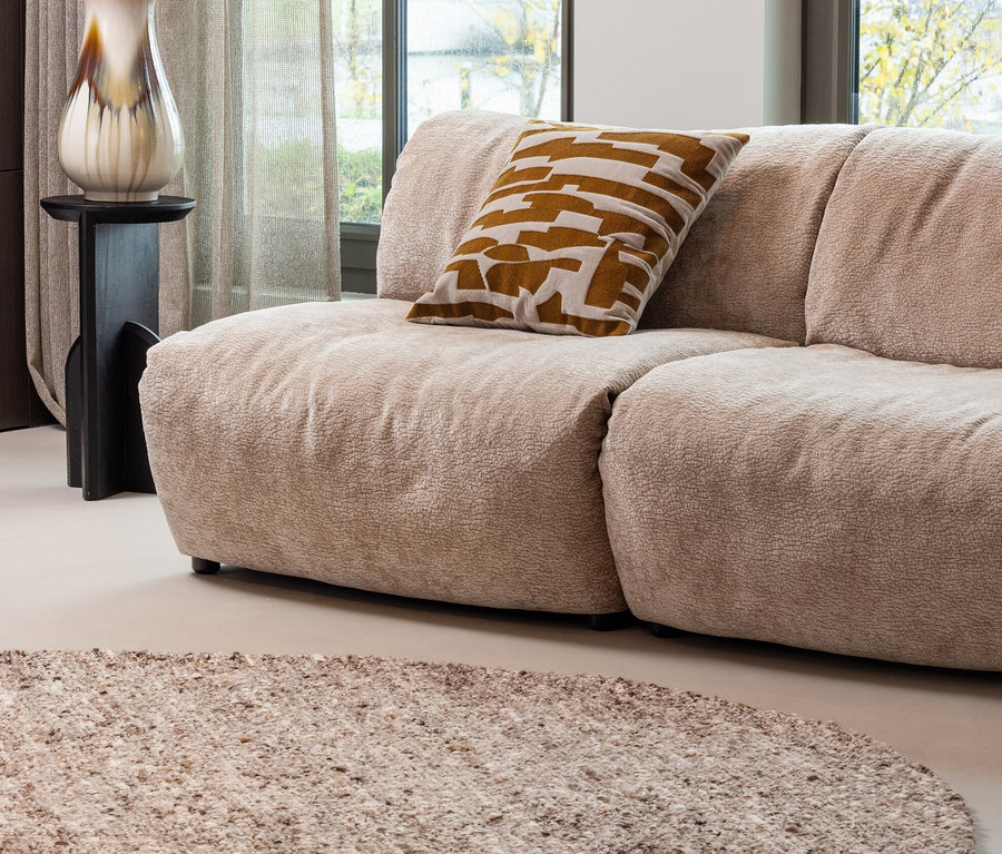 Beiges Sofa mit braun gemustertem Kissen, Vase mit Blumen auf einem Tisch, Fenster und beiger Woood Teppich Efia.