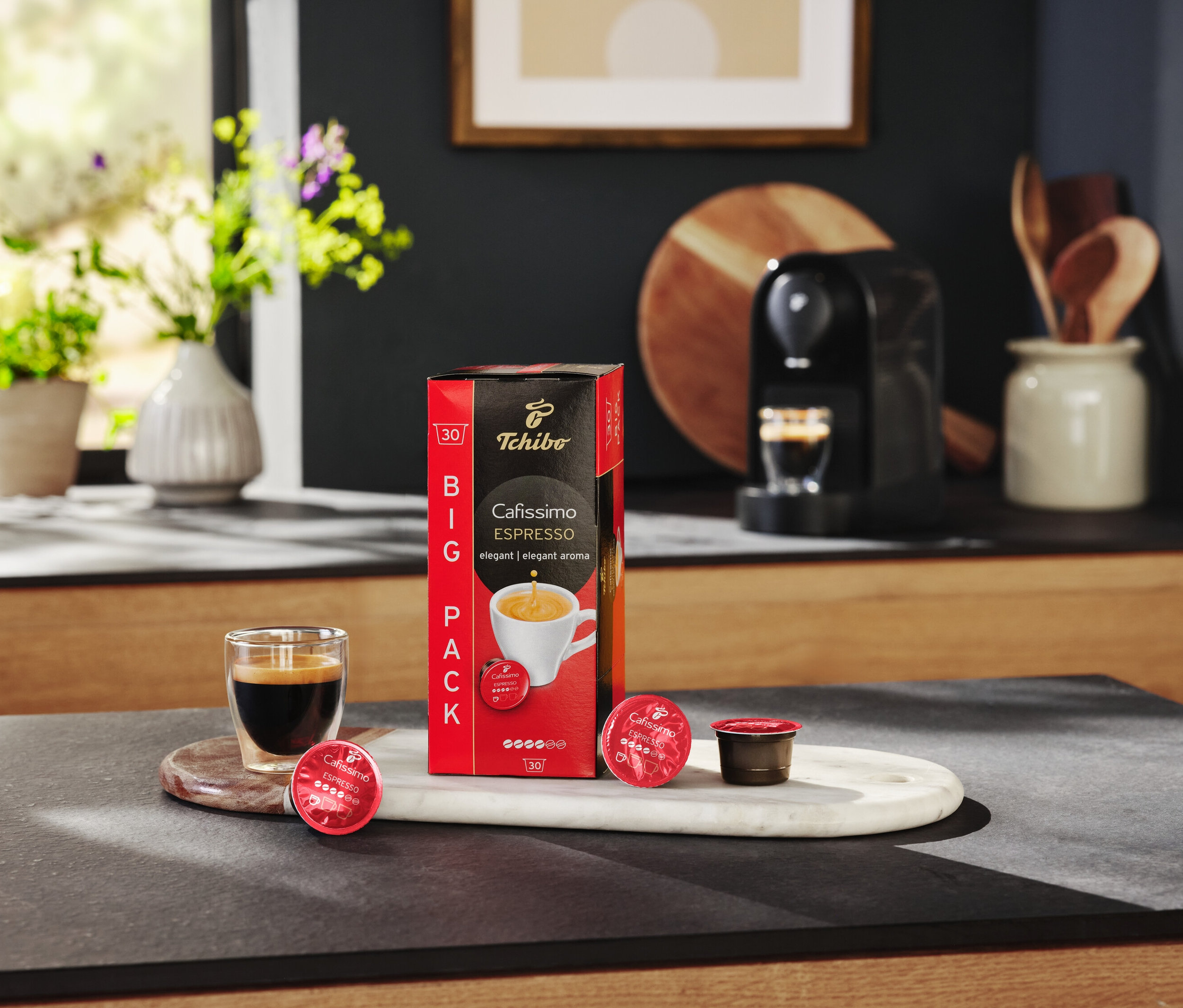 Auf einem Tisch stehen eine Packung Cafissimo Espresso elegant - 10 Kapseln, ein Glas Espresso und eine Kaffeemaschine.