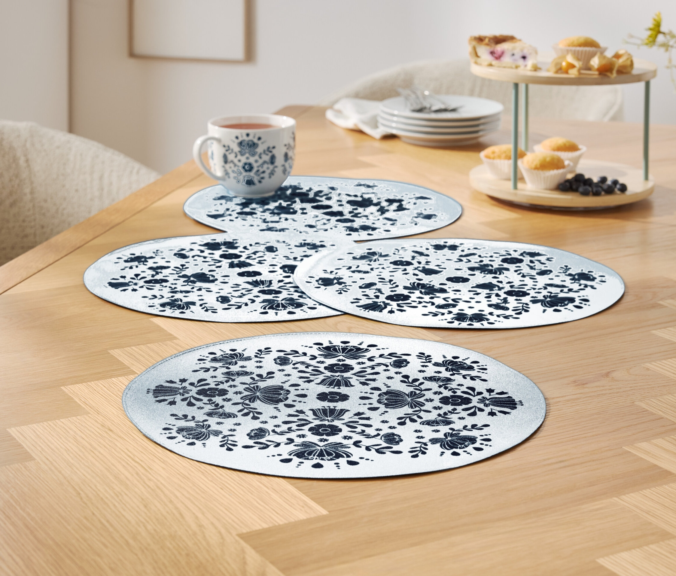 Vier weiße Tischsets mit blauem Blumenmuster auf einem Holztisch, eine Tasse und ein Serviertablett mit Muffins im Hintergrund.